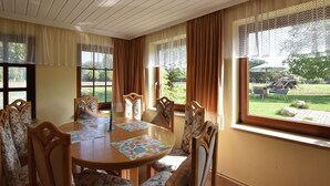 Ein Essbereich mit Holztisch und floralen Stühlen, großen Fenstern mit Vorhängen und Blick ins Freie.