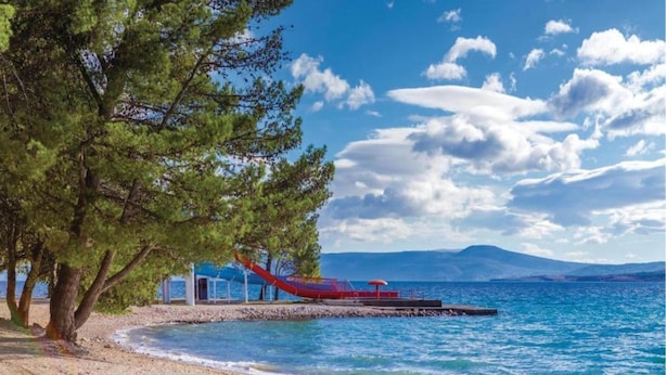 A beach with a wooden pier extending into the sea, a small building with a red roof, and a mountainous backdrop. - Peaceful Villa in Dramalj with Hot Tub (Crikvenica)