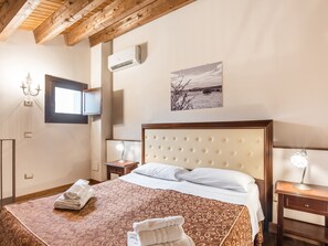 A hotel room with a bed, a bedside table with a lamp, a wall-mounted air conditioner, a picture of a landscape, and a window.