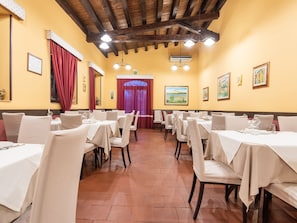 A restaurant with multiple tables and chairs, wooden ceiling beams, and a red curtain entrance.