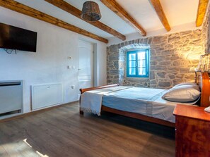A bedroom with a stone wall, a wooden ceiling, a bed, a TV mounted on the wall, and a window with blue shutters. - Gorgeous Villa With Private Swimming Pool and Covered Terrace (Krk)