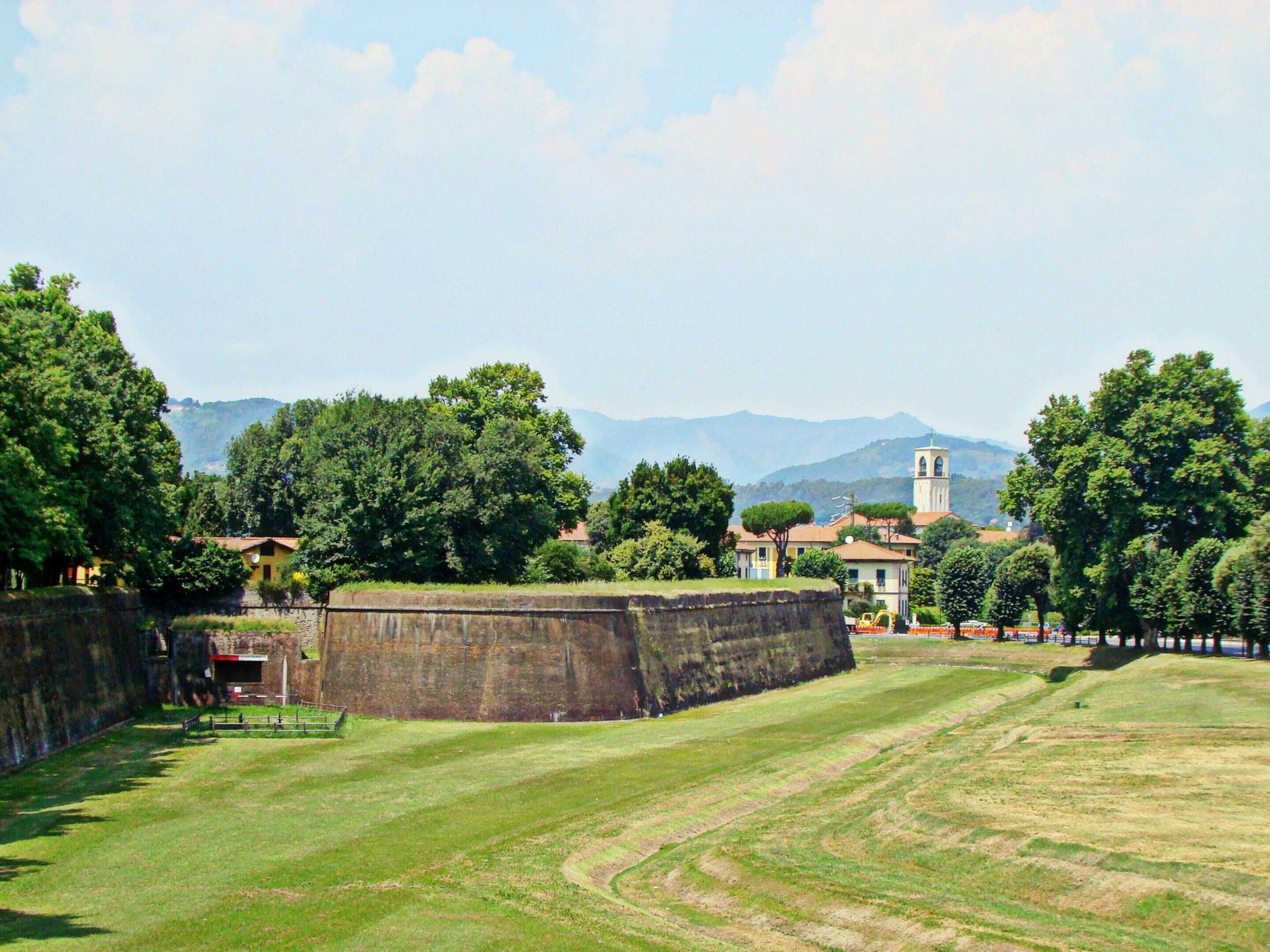 A grassy field with a stone wall and a few buildings in the background.
