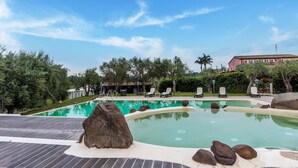 A swimming pool with rocks and lounge chairs, surrounded by trees and a building in the background. - Holiday Home Near Mount Etna Views (Carlentini)
