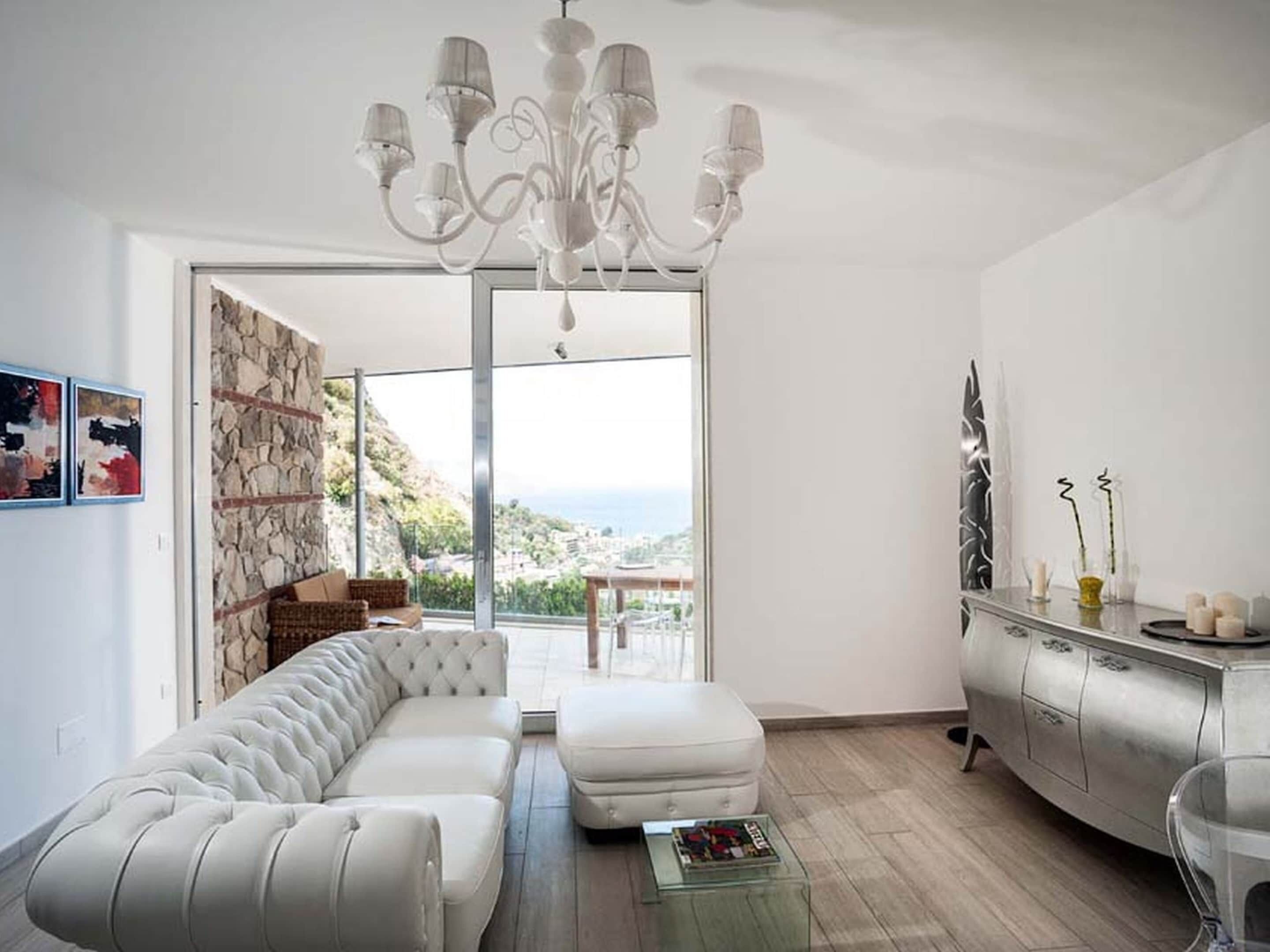 A modern living room with a white sofa, a glass coffee table, and a chandelier. There is a view of the sea through large windows.