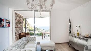 A modern living room with a white sofa, a glass coffee table, and a chandelier. There is a view of the sea through large windows.
