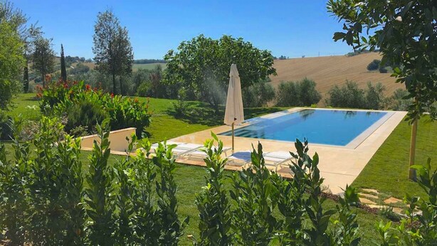 A rectangular swimming pool with a white umbrella, surrounded by a well-maintained lawn and various plants. - Belvilla by OYO Villa Colle (Recanati)