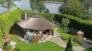 A thatched-roof gazebo with a stone wall, surrounded by a well-maintained garden and a lake view. - Holiday Home by Lake in Nowa Wioska (Lubrza)