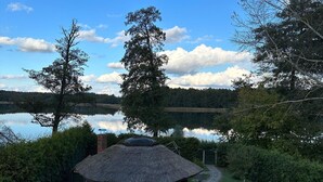 A thatched-roof hut with a stone wall, a stone pathway, and a view of a lake surrounded by trees. - Holiday Home by Lake in Nowa Wioska (Lubrza)