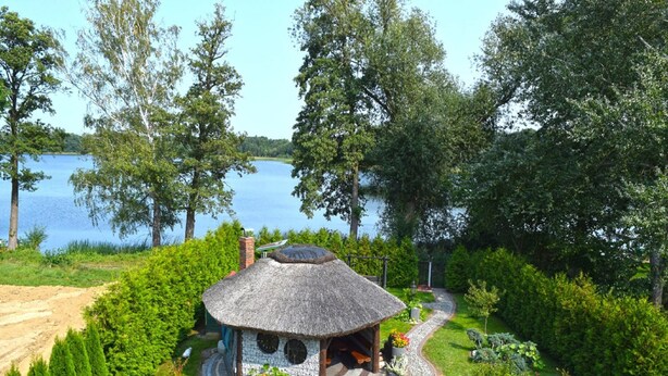 A thatched-roof cabin surrounded by lush greenery and a winding stone path. - Holiday Home by Lake in Nowa Wioska (Lubrza)