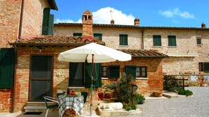 Un edificio rustico con facciata in mattoni, tetto in tegole e un piccolo balcone. C'è un tavolo con tovaglia e sedie all'esterno, e una pianta in vaso nelle vicinanze.