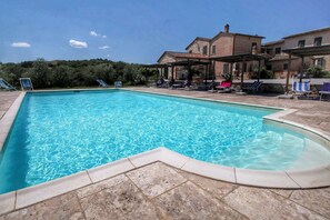 Une piscine rectangulaire à l’eau d’un bleu limpide, entourée de carreaux de pierre. Des chaises longues et des parasols sont disposés autour de la piscine.