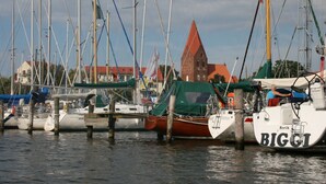 A marina with multiple sailboats docked, a church with a red roof, and a clear sky. - Ferienhaus mit Balkon und Garten in Zweedorf (Bastorf)