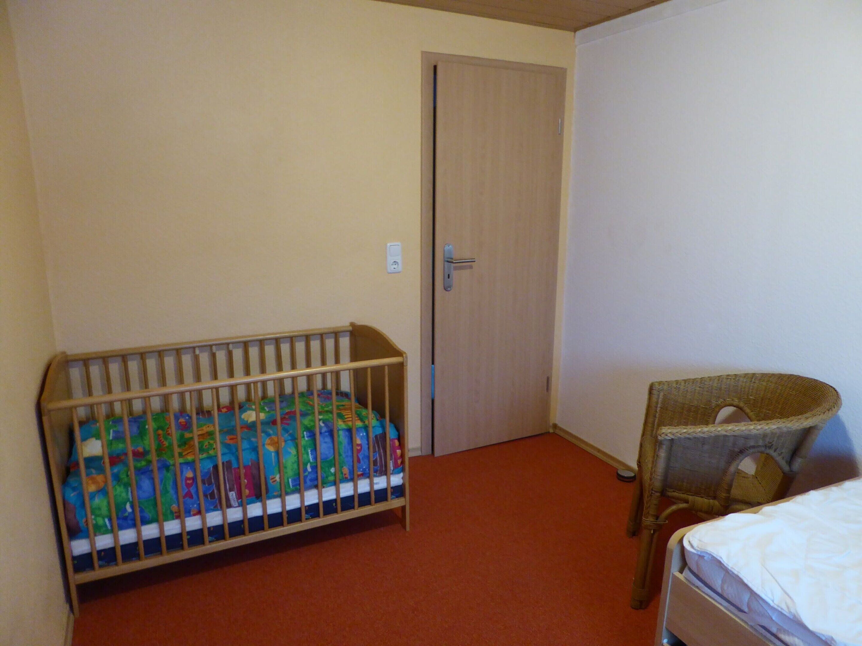 Een kamer met een ledikant, een rieten stoel en een gesloten deur.