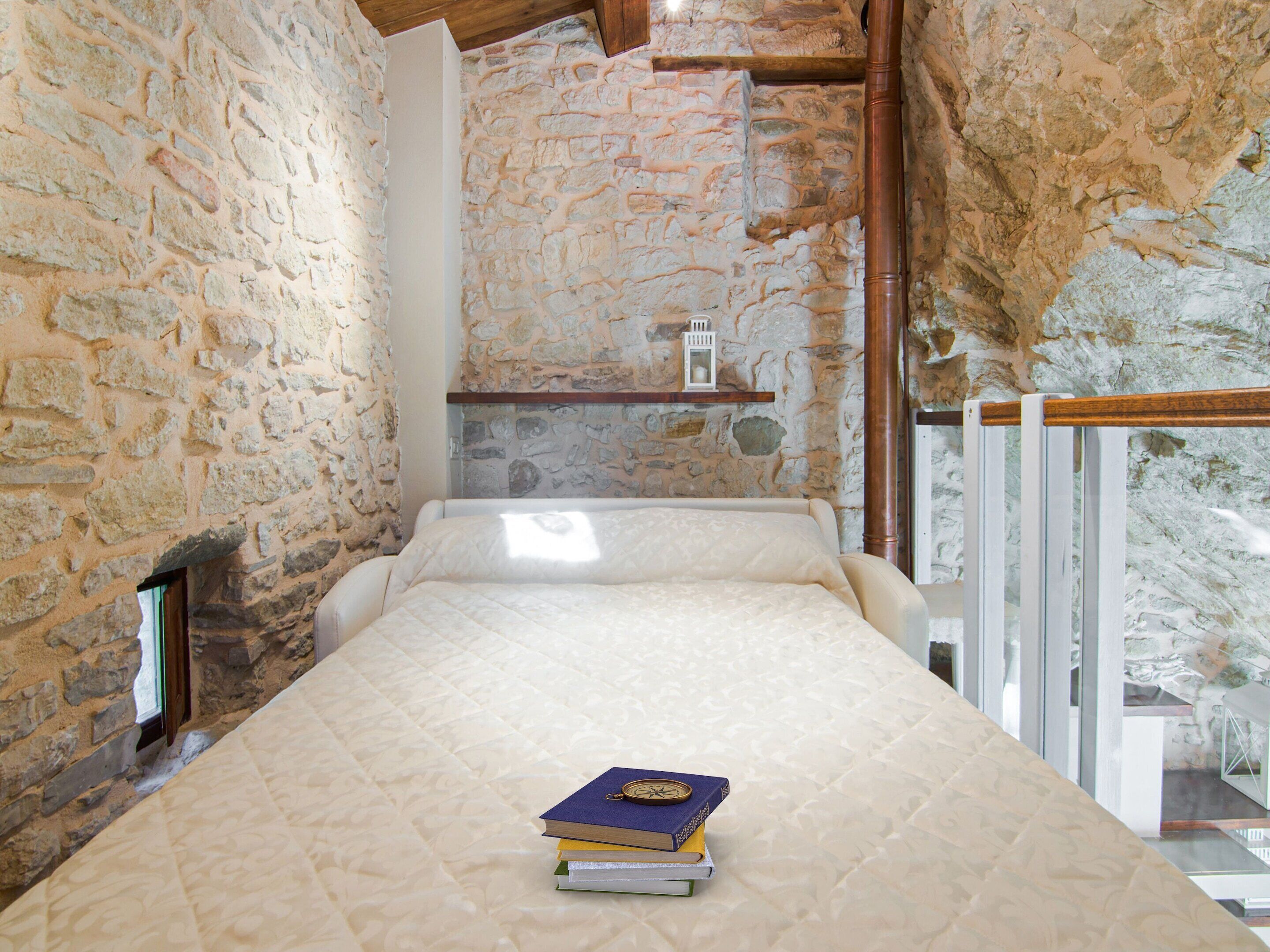Una habitación estrecha, con paredes de piedra, una cama y un pequeño estante con libros y un farol.