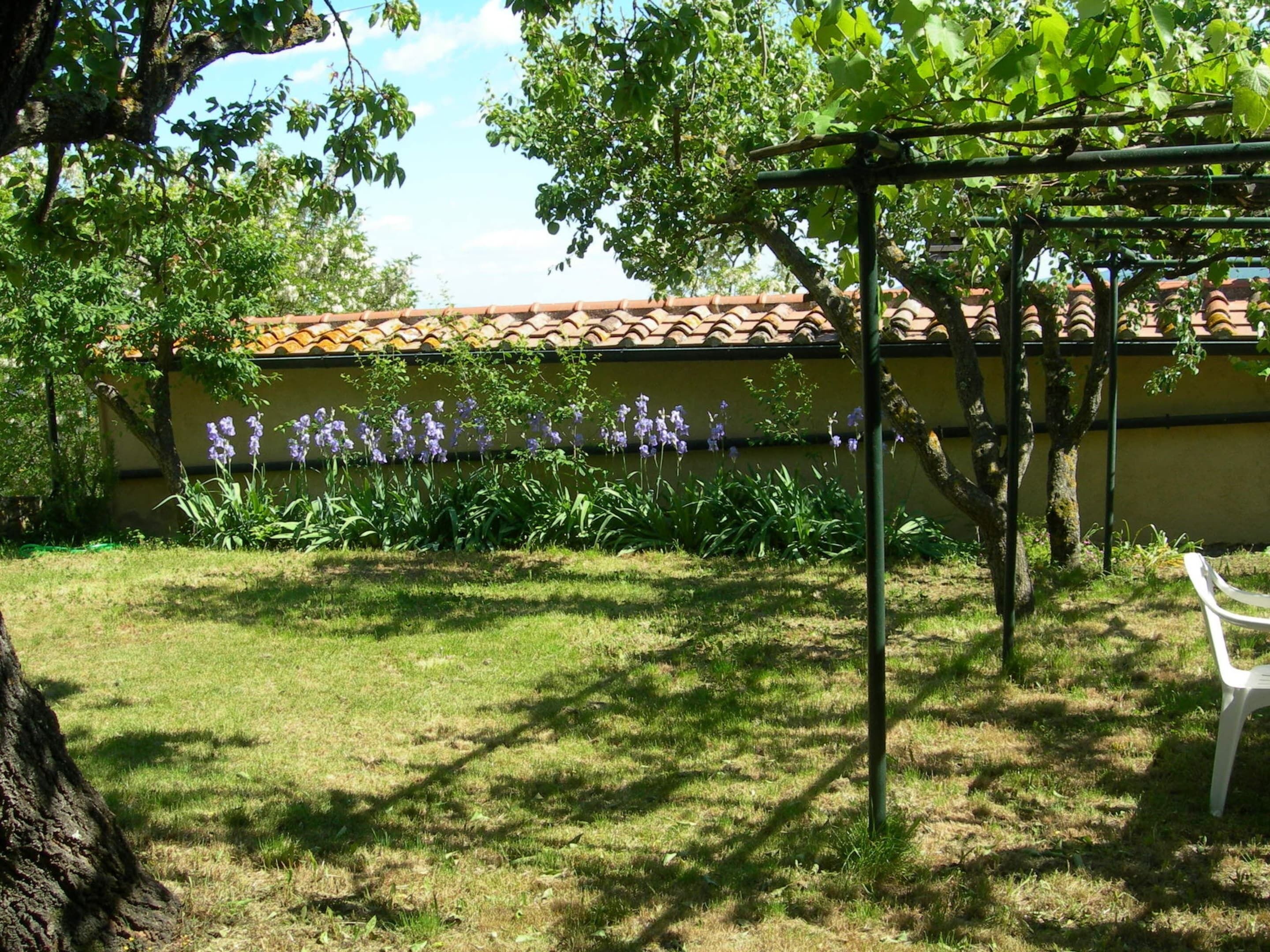 A garden with a pergola, a white plastic chair, and a building with a tiled roof in the background.
