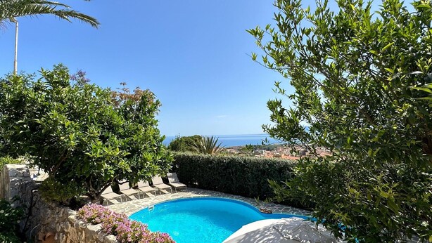 Una piscina con un muro de piedra, rodeada de follaje y vista al mar.