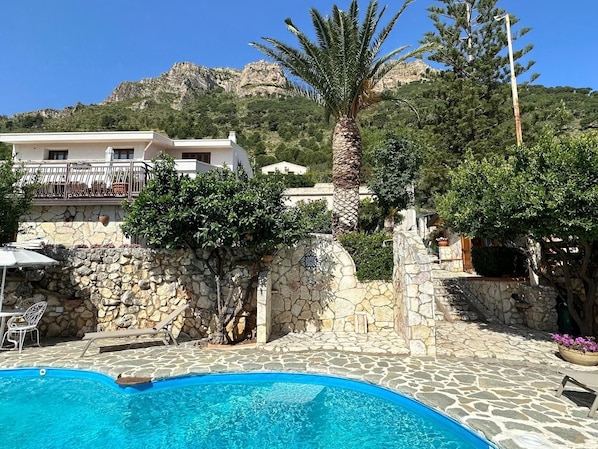 A pool area with stone flooring, a palm tree, and a building with a balcony in the background. - Spacious Apartment With Communal Pool Near the Sea (Castellammare del Golfo)
