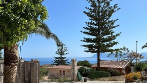 Un patio empedrado con vistas al mar, rodeado de árboles y un pequeño edificio con techo de tejas.