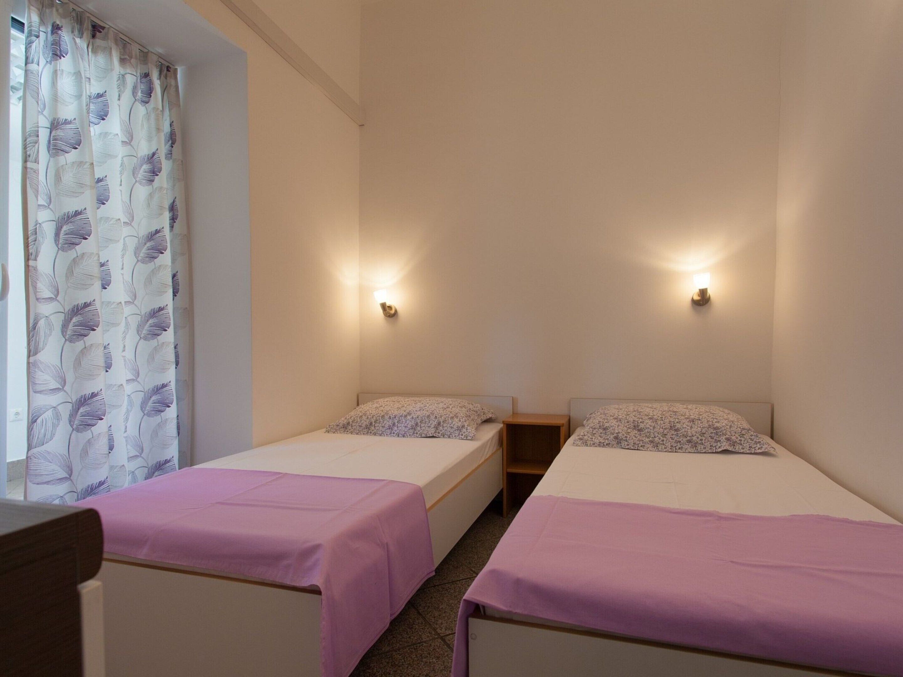A hotel room with two single beds, a small wooden nightstand, and a window with a floral curtain.