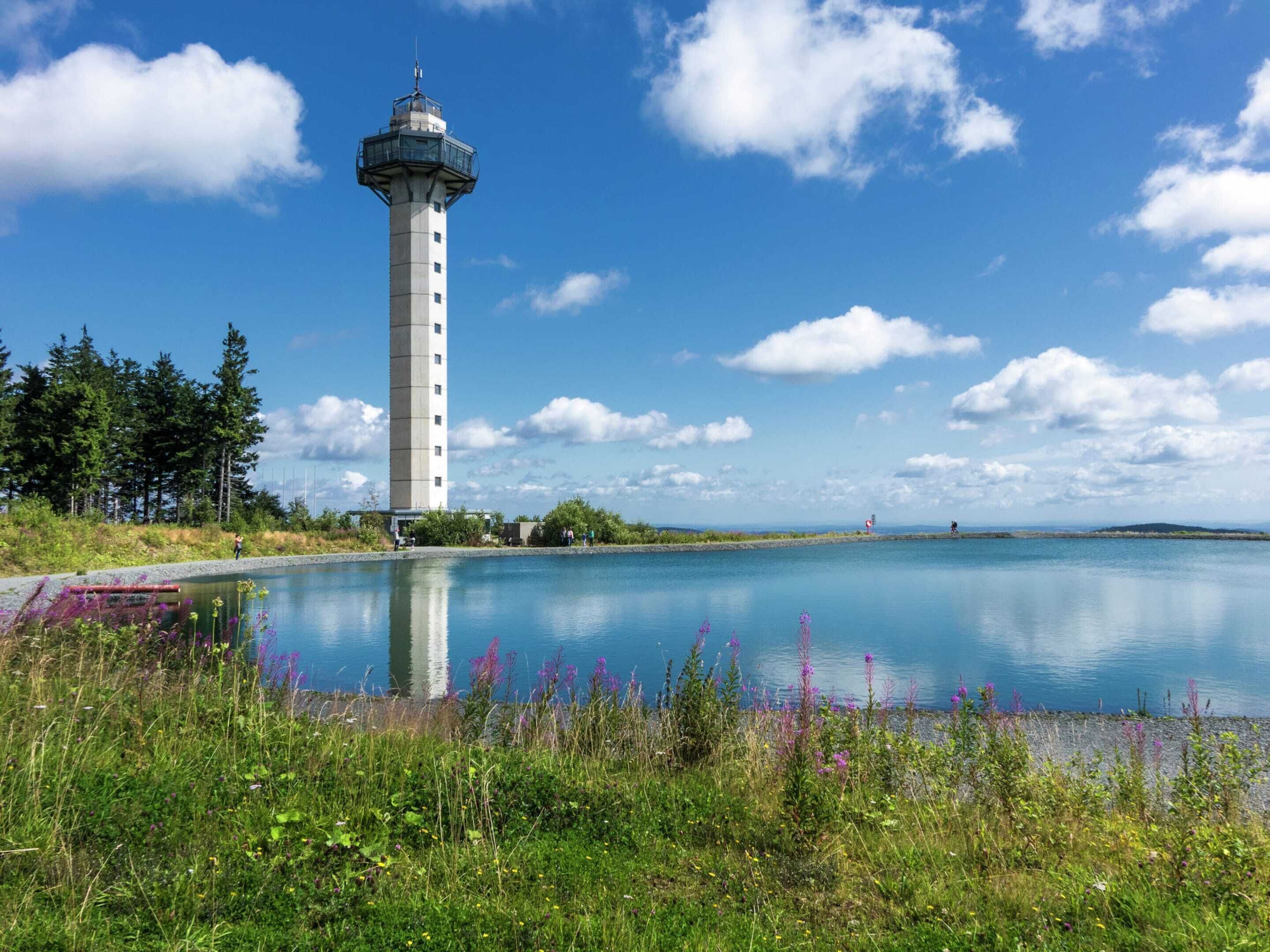 Un phare au bord d’un lac paisible, entouré de verdure et sous un ciel dégagé.