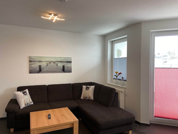 A modern living room with a dark brown sectional sofa, a wooden coffee table, a painting of a pier, a window with a view of a red building, and a ceiling light fixture.