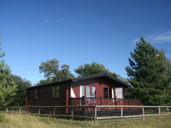 Une cabane rouge avec une véranda entourée d’une clôture, au milieu des arbres et de l’herbe.