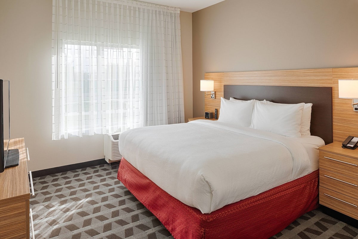 A hotel room with a large bed, a bedside table with a lamp, a TV, and a window with sheer curtains.