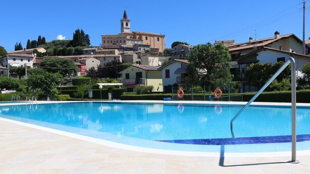 Una piscina con un bordeado de baldosas, un flotador salvavidas y una escalera metálica. Al fondo, se ven casas, una iglesia y una colina con árboles.