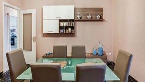 A dining area with a glass table, four chairs, a chandelier, a white cabinet with decorative items, and a wooden shelf above.
