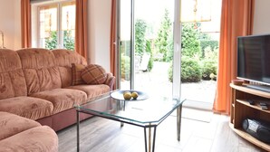 Een woonkamer met een bank, een glazen salontafel, een televisie en een schuifpui die uitkomt op een tuin.