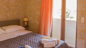 A bedroom with a stone wall, a bed with white and gray bedding, a lamp on a nightstand, and a balcony with an orange curtain. - Beach View Villa in Bali near City Center with Private Pool (Mylopotamos)