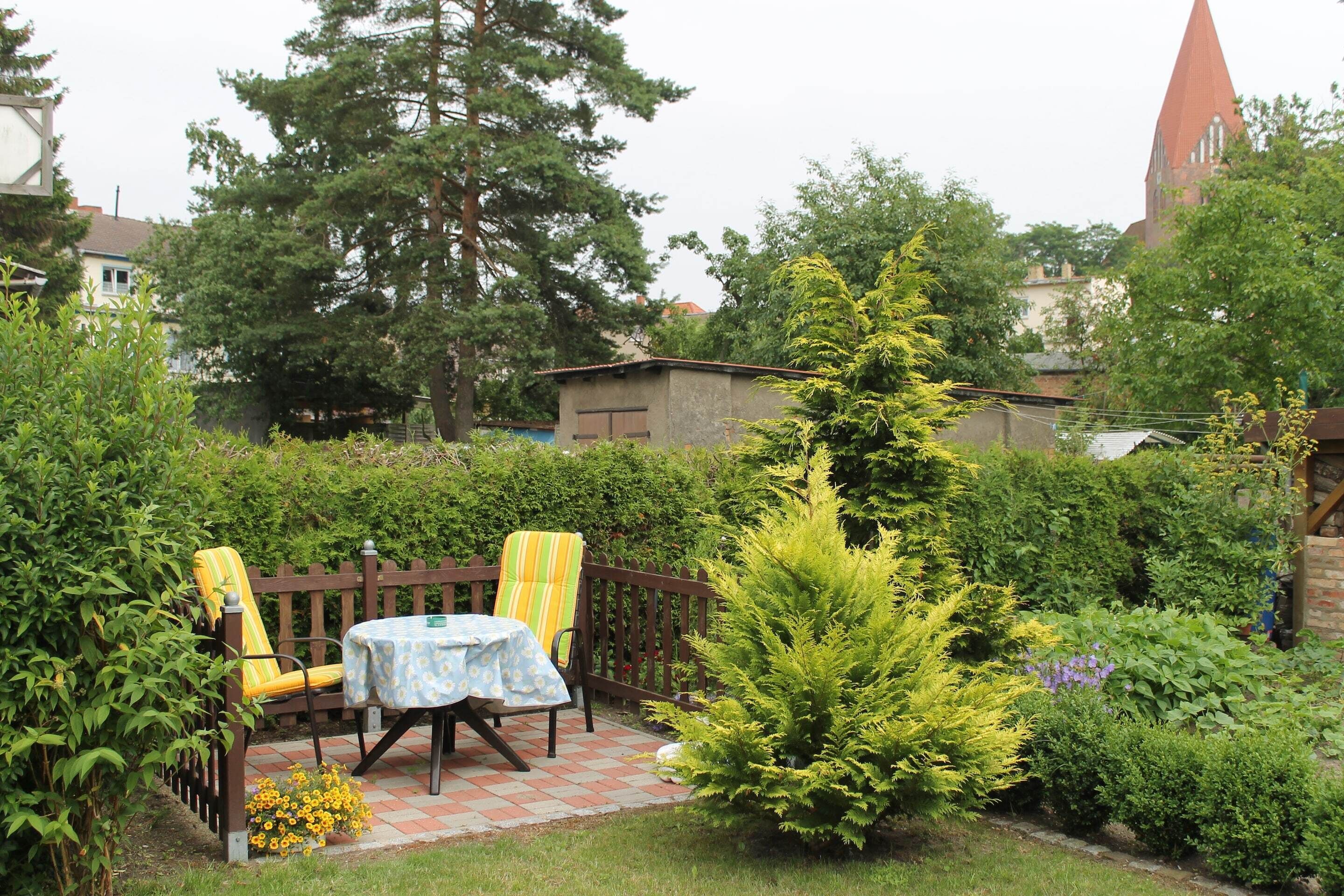 Ein Garten-Terrasse mit Tisch und zwei Stühlen, umgeben von Pflanzen und einem Holzzaun.