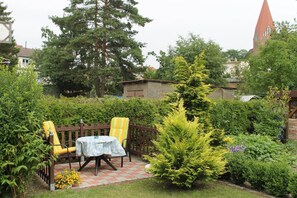 Une terrasse de jardin avec une table et deux chaises, entourée de différentes plantes et d’une clôture en bois.