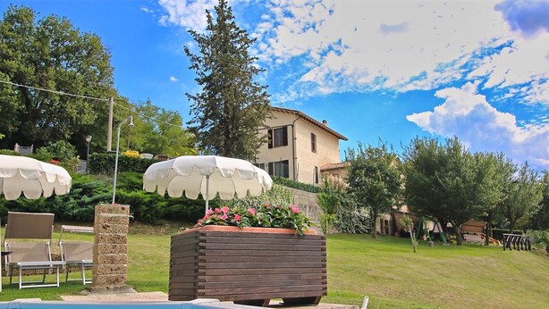 A swimming pool with a tiled edge, a wooden planter with flowers, outdoor chairs, and a large umbrella. - Flat in Piticchio With Scenic Views & Pool (Arcevia)