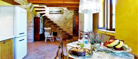 A rustic kitchen with a wooden ceiling, a white refrigerator, and a dining table set with fruits and a vase.