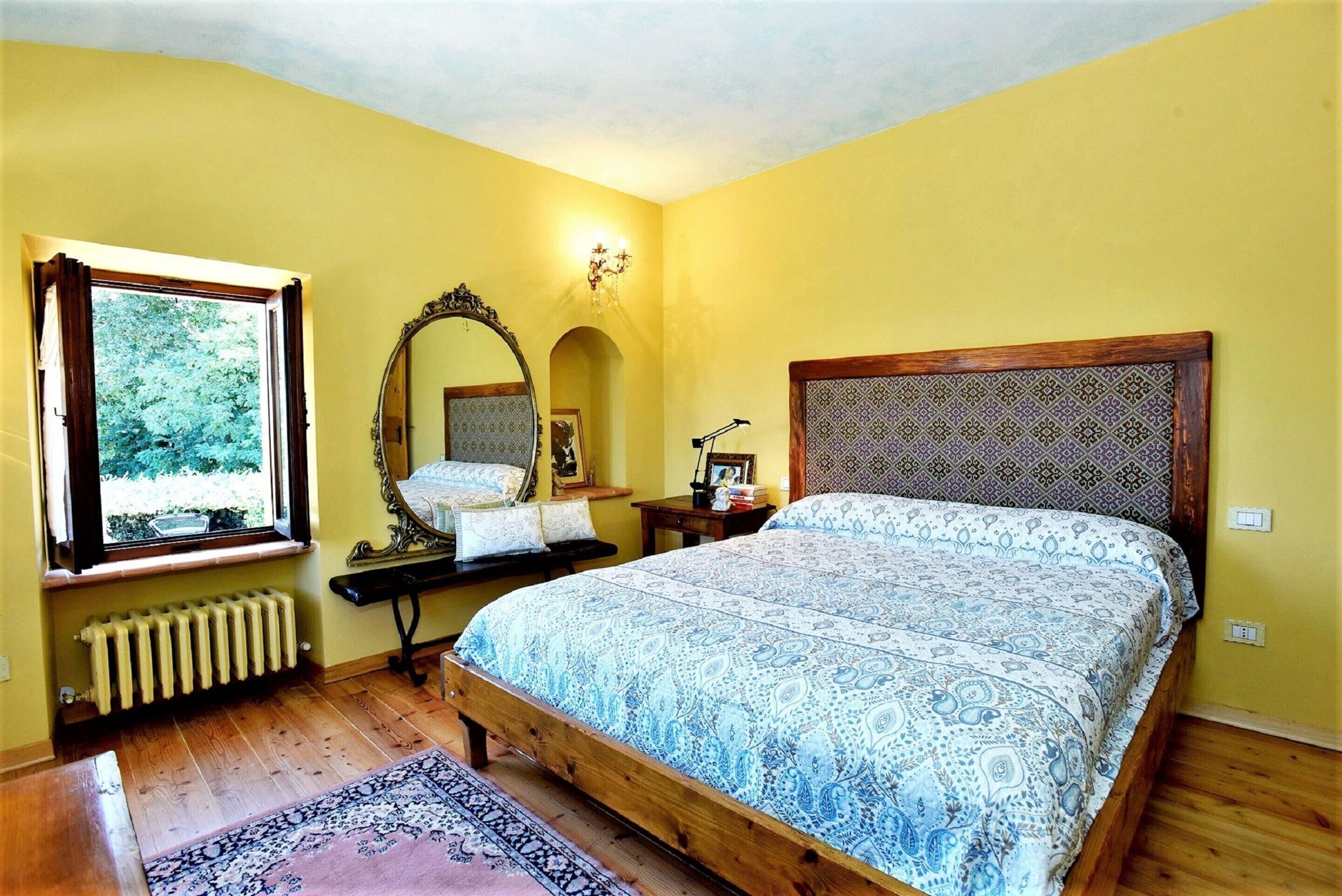 Quarto com cama, janela, radiador, espelho e mesa de cabeceira.
