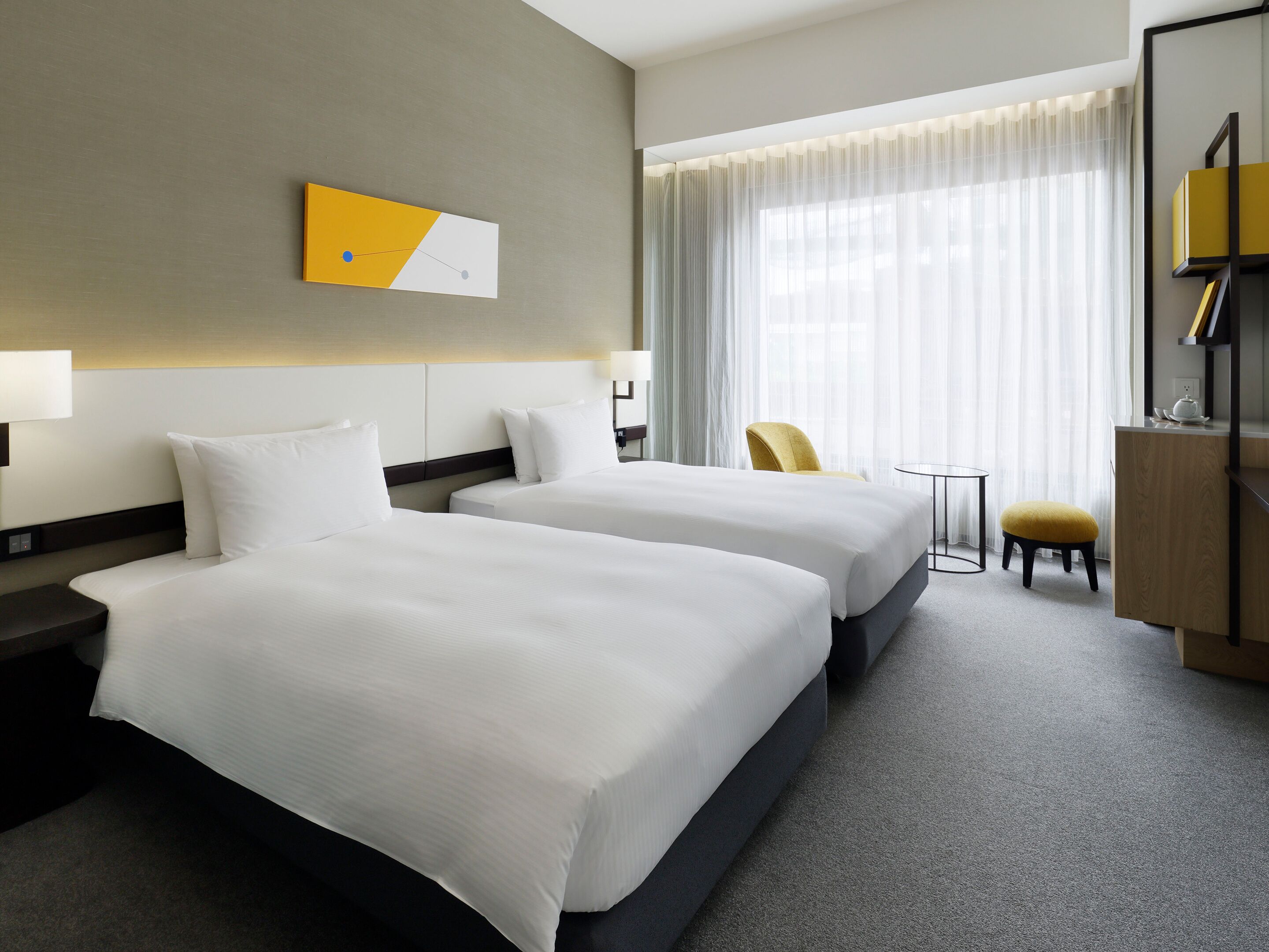 Een hotelkamer met twee bedden, een bureau, een stoel en een groot raam met dunne gordijnen.