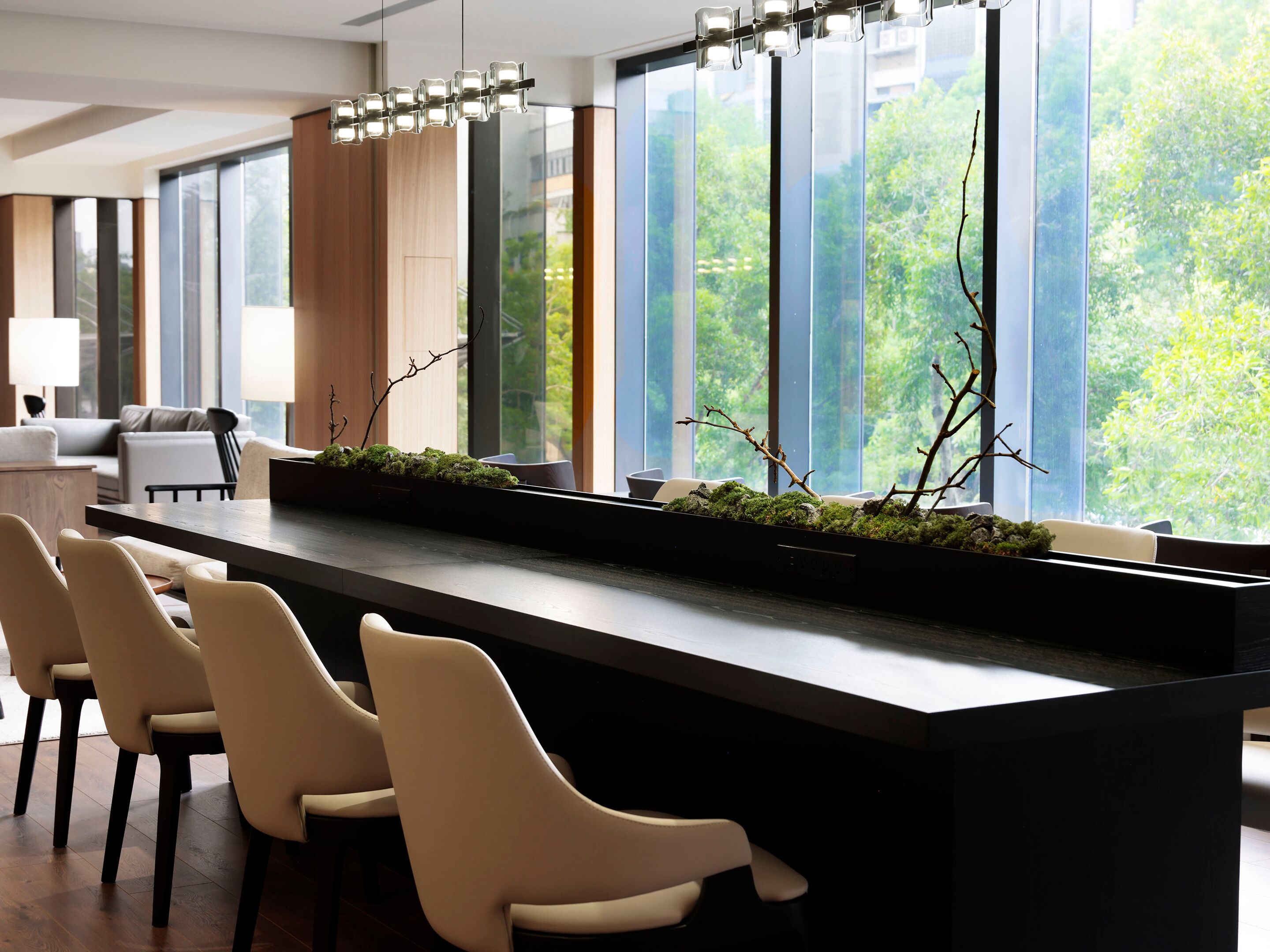 A modern bar area with a long counter, several chairs, and a view of greenery through large windows.