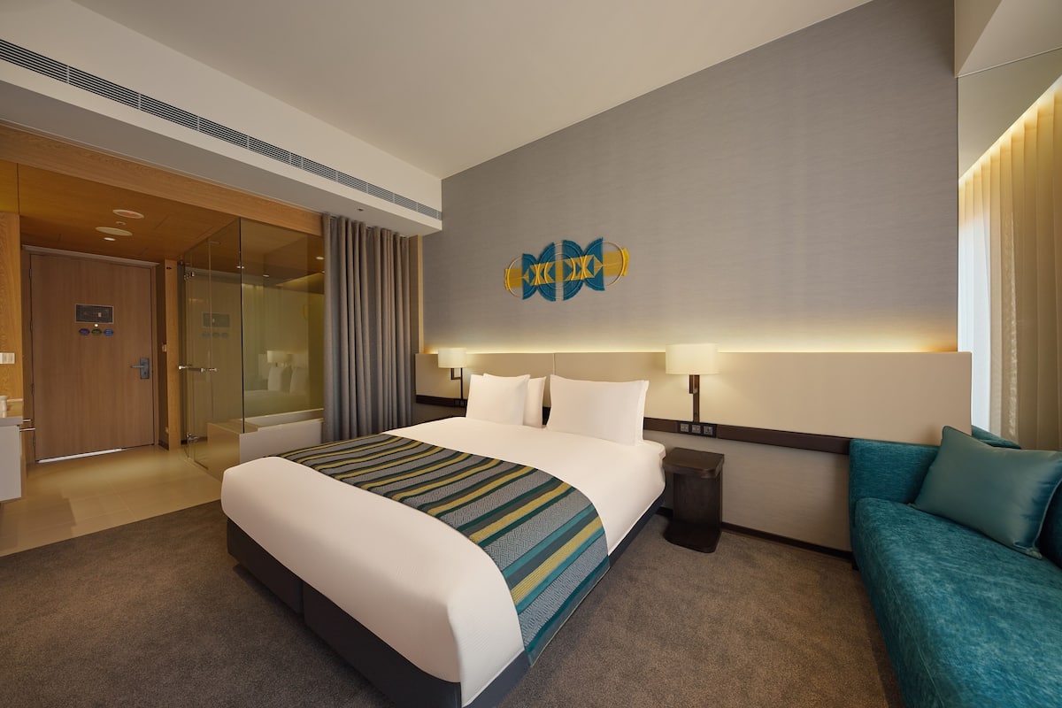 A modern hotel room with a large bed, a sofa, a glass shower enclosure, and a wall-mounted artwork.