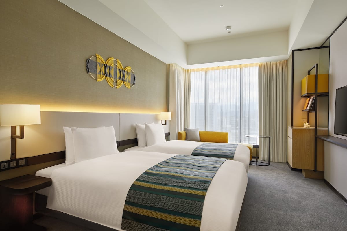 A hotel room with two beds, a desk, a television, and a large window with curtains.