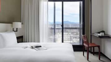 A hotel room with a large bed, a desk, a chair, and a view of the cityscape through a large window.