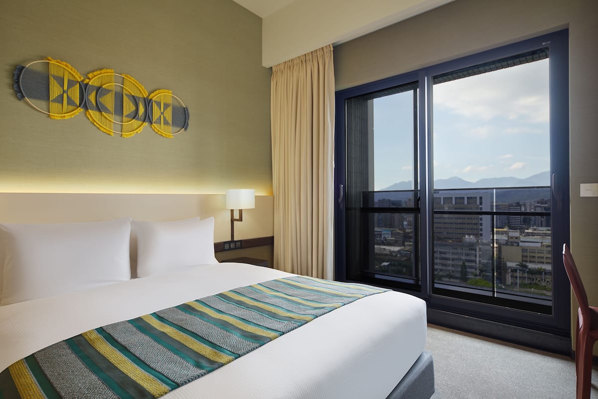 A hotel room with a large bed, a view of the city through a window, and a decorative wall piece.