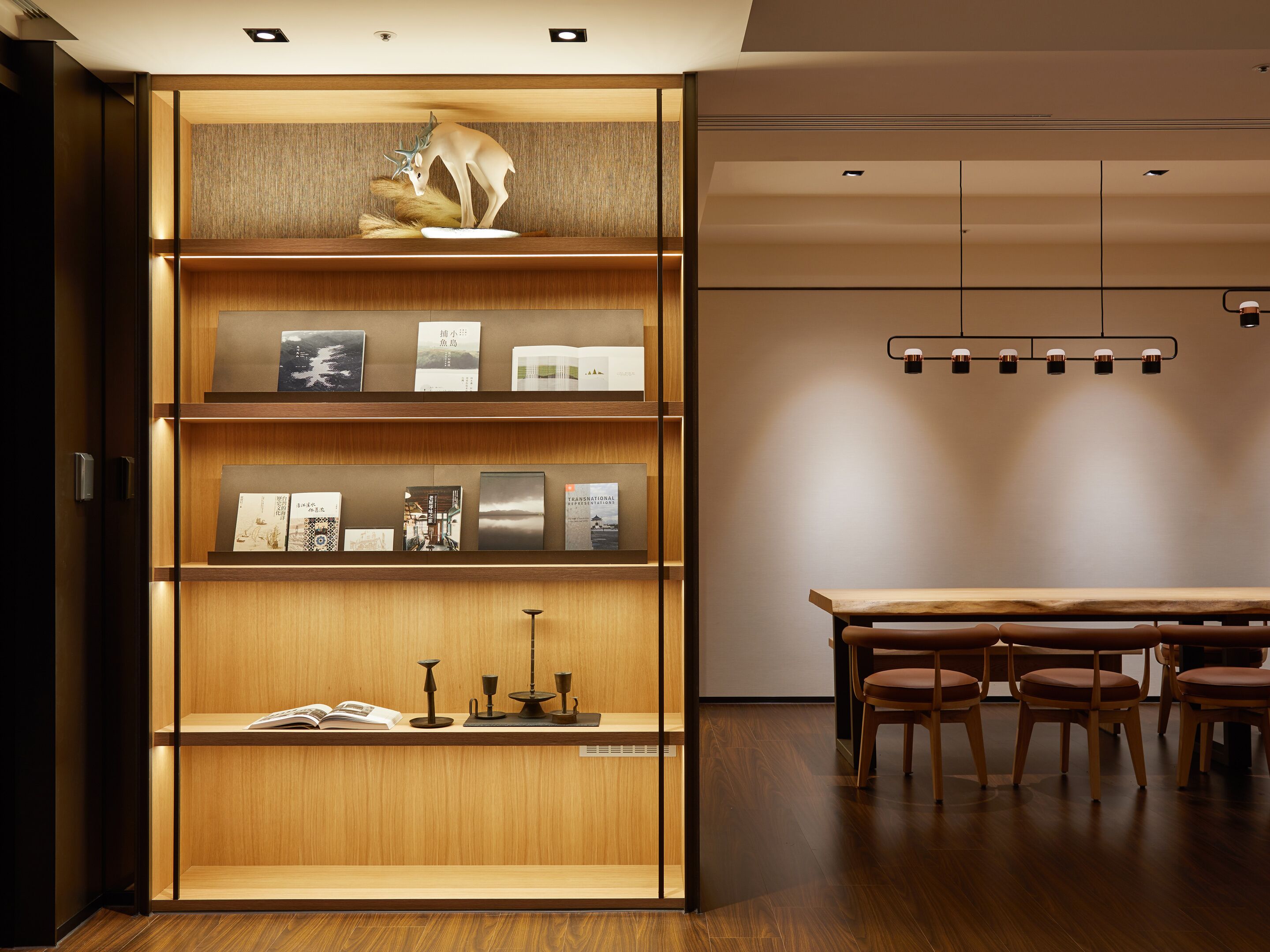 A modern interior with a wooden bookshelf displaying books and decorative items, a dining area with wooden tables and chairs, and recessed ceiling lighting.