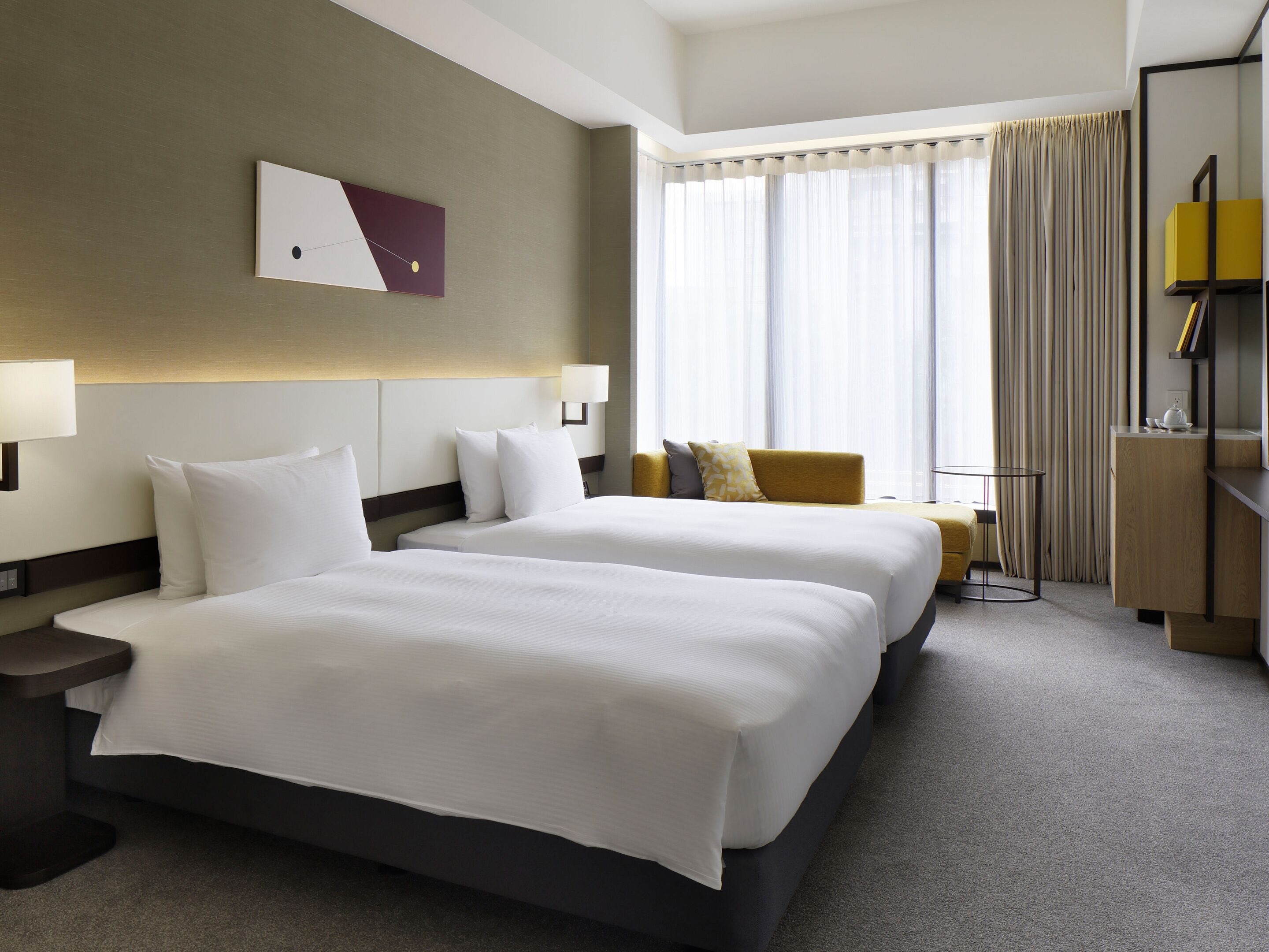 Habitación de hotel con dos camas, una mesita de noche, una lámpara, un sofá, una pequeña mesa y una estantería.