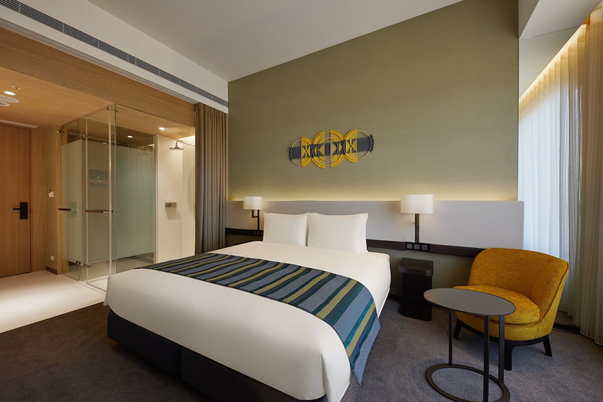 A hotel room with a large bed, a glass shower enclosure, a small table, and a yellow armchair.