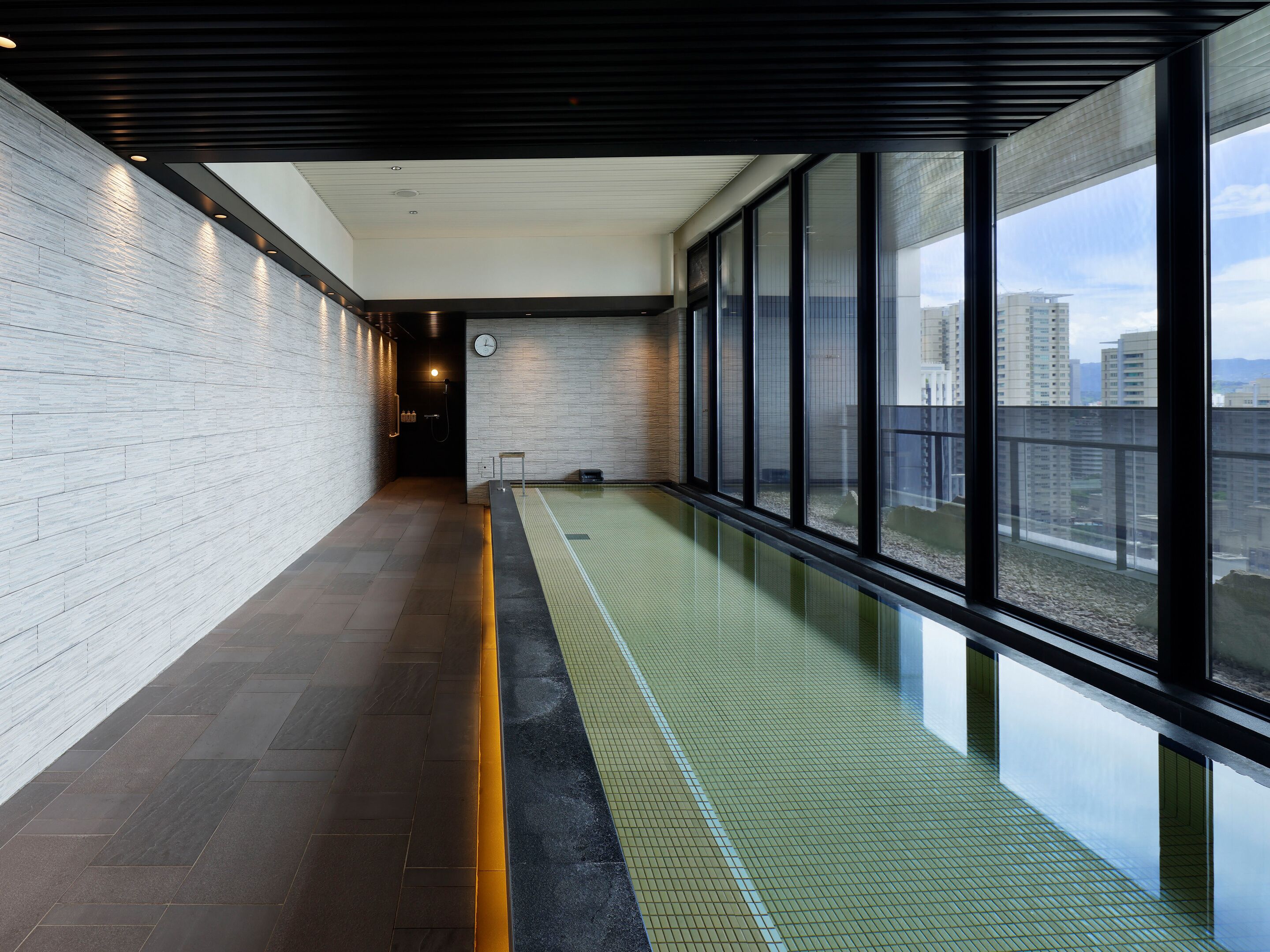 A modern indoor swimming pool with a tiled floor, large windows, and a clock on the wall.
