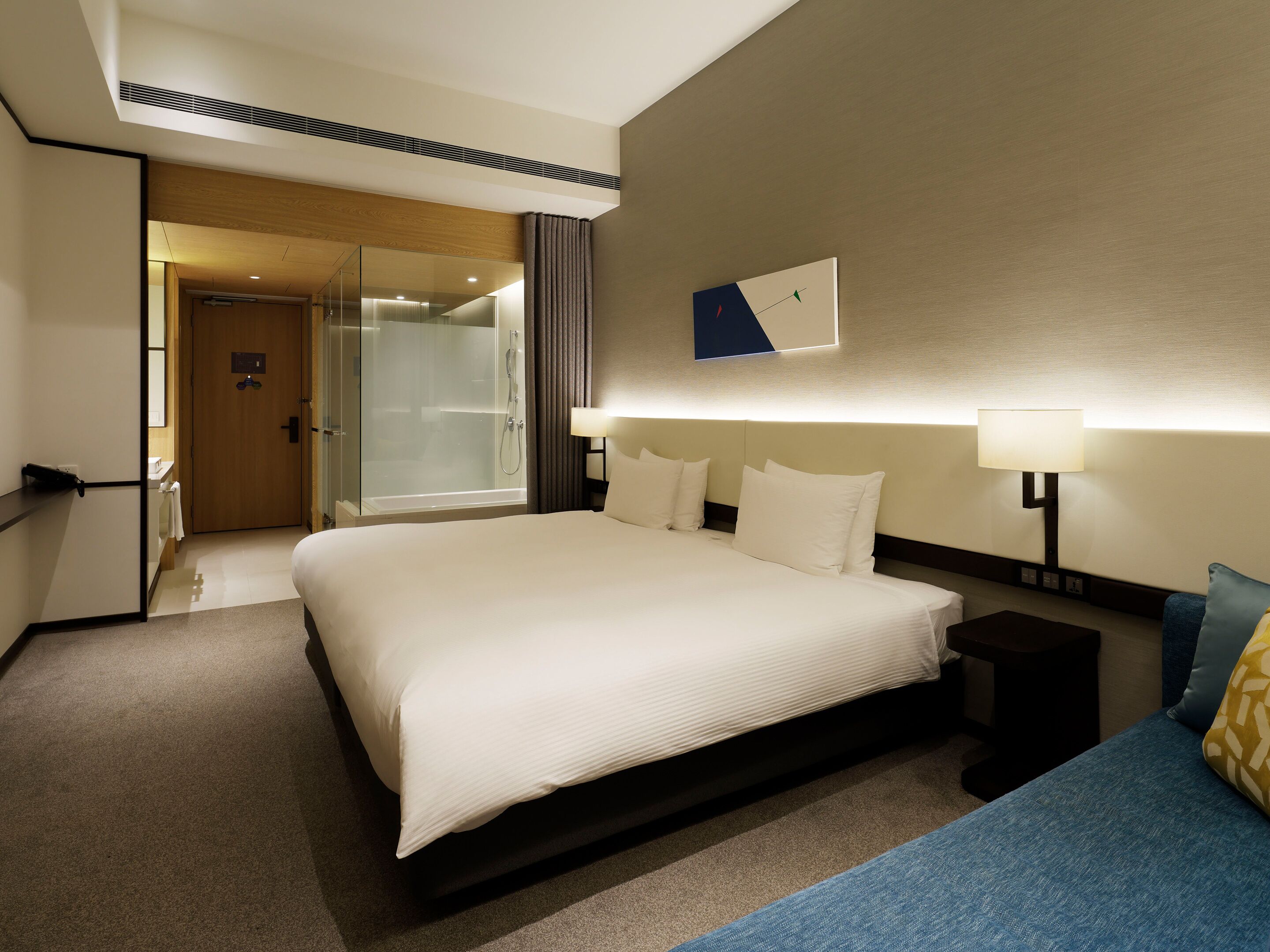 Habitación de hotel con una cama grande, lámparas de noche, cabina de ducha de vidrio y vistas al pasillo.