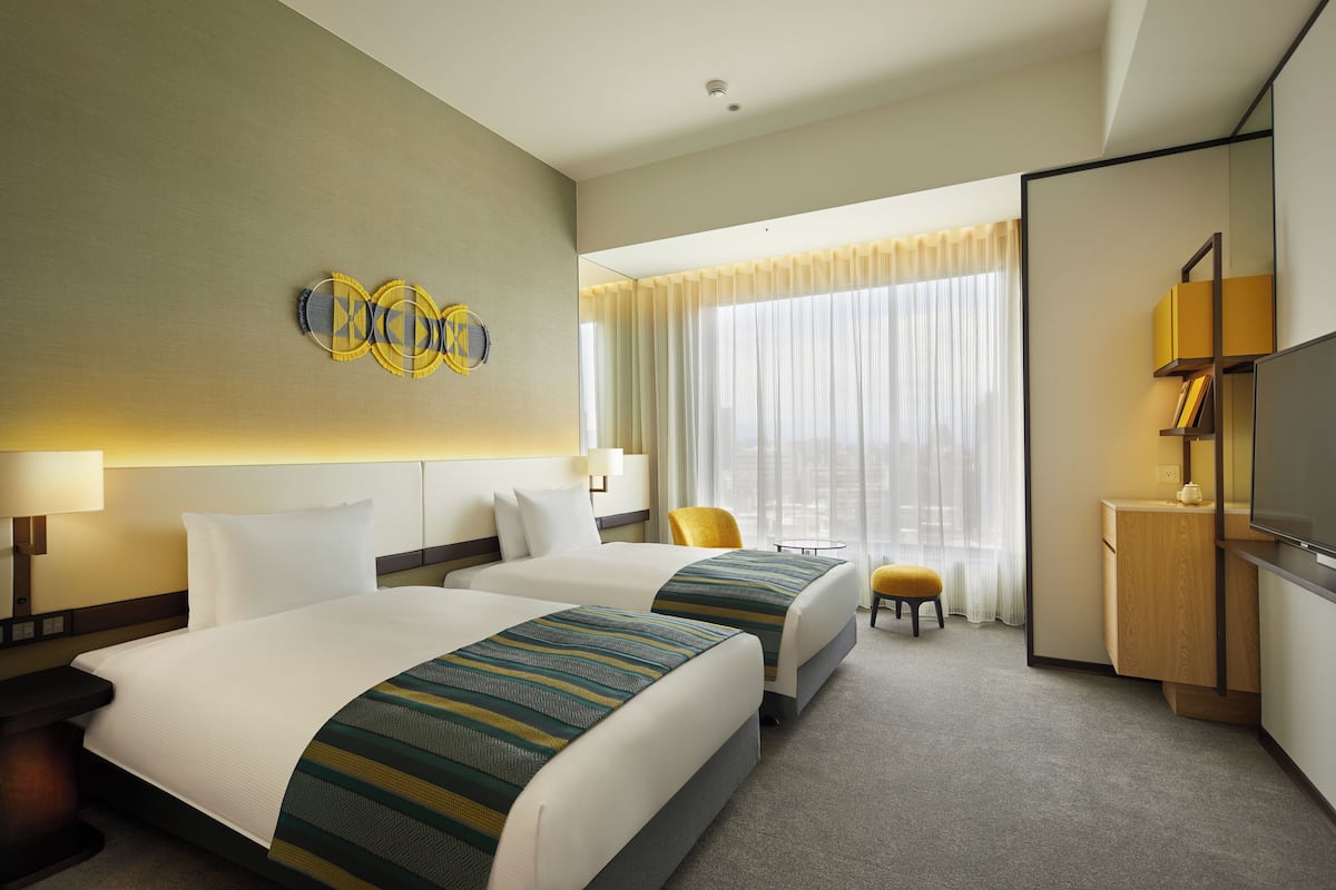 A hotel room with two beds, a desk, a chair, a TV, and a large window with curtains.
