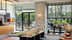 Montaje de bufé con diversos alimentos, una zona de bar con bebidas y un ventanal que ofrece vistas a la vegetación exterior.