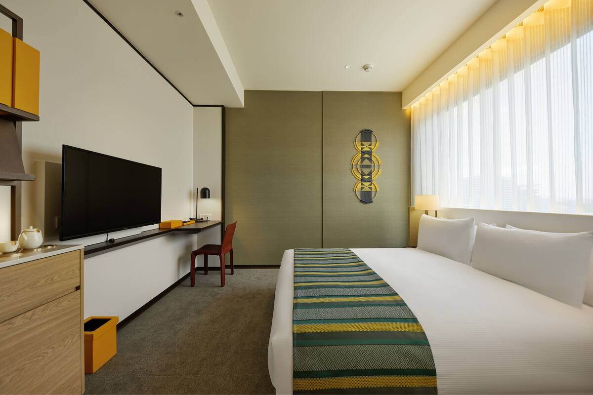 A hotel room with a bed, a desk with a flat-screen TV, a chair, and a wall-mounted artwork.